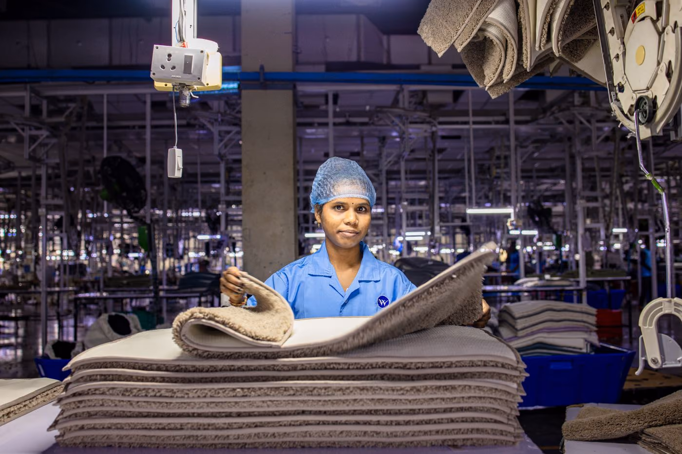 Operadora de produção em linha de fábrica, de uniforme e touca, manuseando material.